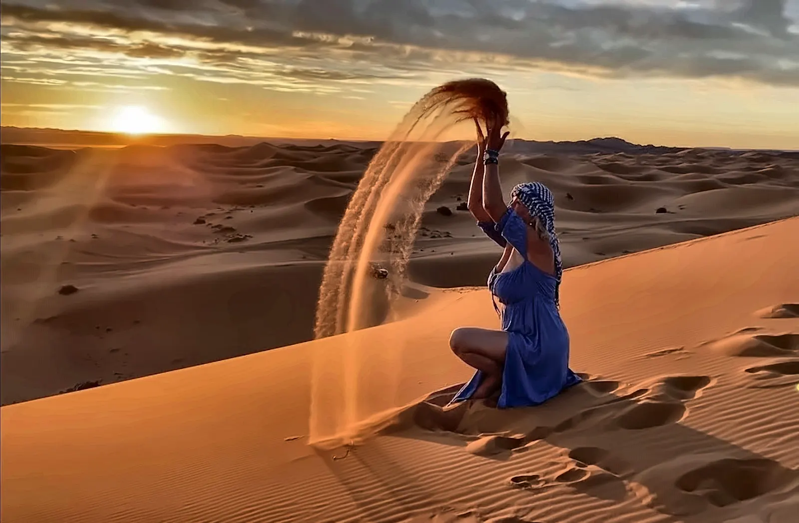 Una viaggiatrice in abito blu lancia la sabbia in aria sulle dune dell’Erg Chebbi, al tramonto